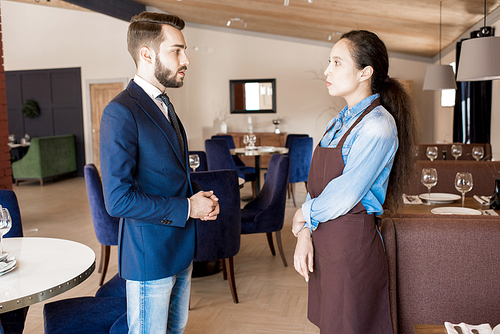 Serious displeased handsome restaurant manager in smart casual outfit talking to young waitress in apron while explaining her duties in modern establishment