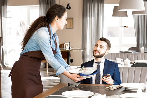 Content handsome young businessman having lunch in restaurant: bearded man satisfied with restaurant service looking at waitress putting plate on table