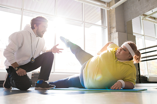 Full length portrait of obese young woman stretching while working out with personal coach in sunlit fitness club