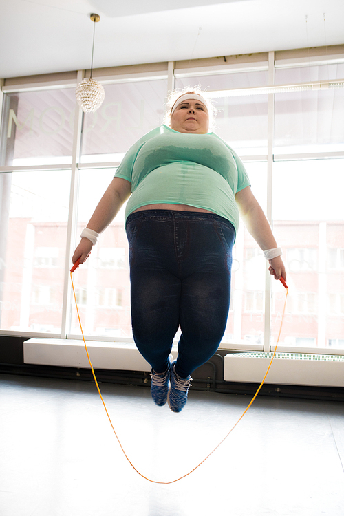 Full length portrait of obese young woman jumping rope during workout in fitness studio