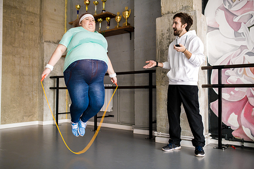 Full length portrait of fat young woman jumping rope during workout with fitness coach, copy space