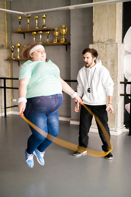 Full length portrait of angry coach shouting at  fat woman jumping rope during training in fitness club