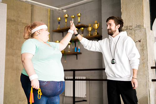 Waist up portrait of fitness coach high fiving with fat young woman during workout