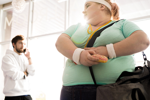 Low angle Waist up portrait of obese young woman holding donut leaving fitness training, copy space