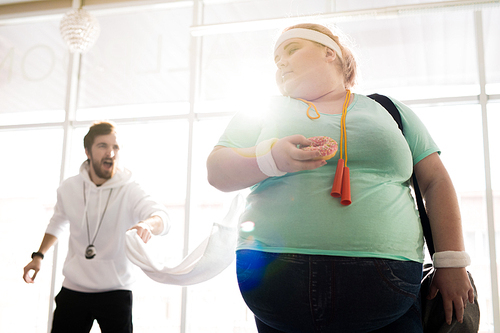 Waist up portrait of obese young woman holding donut while leaving fitness training with angry coach in background, copy space