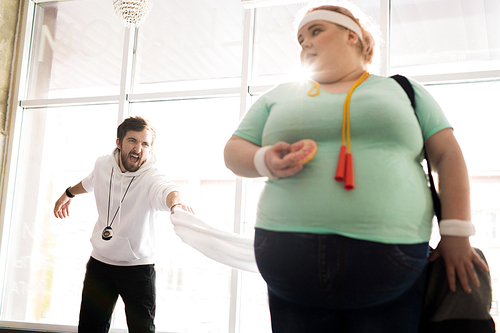 Portrait of obese young woman holding donut while leaving fitness training with angry coach in background, copy space