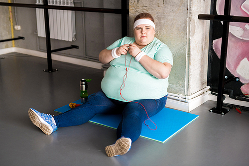 Full length portrait of pensive fat woman sitting on mat while taking break in workout, copy space