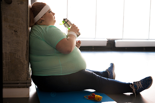 Full length side view portrait of pensive fat woman sitting on mat and drinking water while taking break in workout