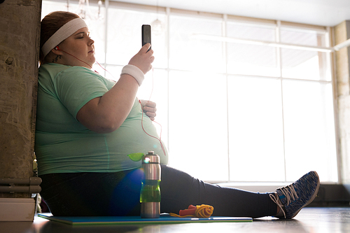 Full length side view portrait of pensive fat woman sitting on mat and using smartphone while taking break in workout