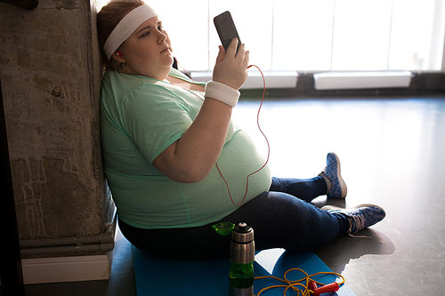 Full length portrait of pensive fat woman sitting on mat and using smartphone while taking break in workout