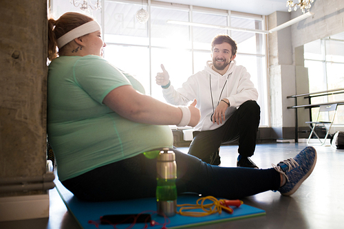 Full length portrait of obese young woman sitting on mat and talking to coach while taking break in workout