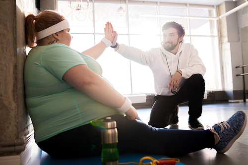 Full length portrait of obese young woman sitting on mat and doing high five with coach while taking break in workout