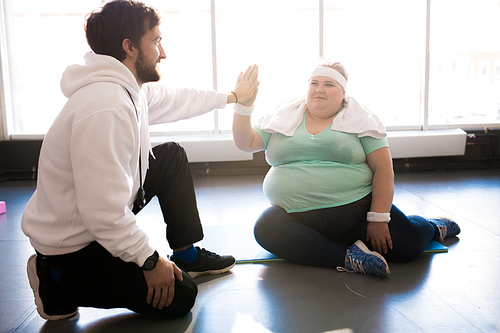 Full length portrait of obese young woman sitting on floor and doing high five with coach while taking break in workout