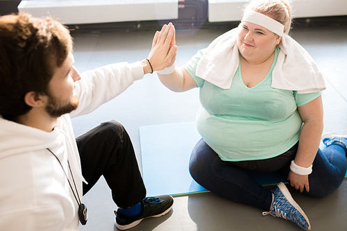 High angle  portrait of smiling obese young woman sitting on floor and doing high five with coach while taking break in workout