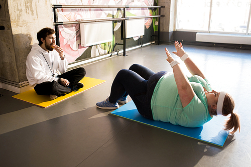 Full length portrait of obese woman doing crunches during workout in fitness club with coach watching her