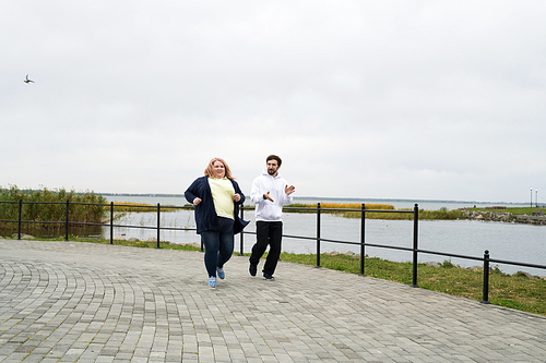 Full length portrait of overweight woman jogging outdoors with personal trainer, copy space