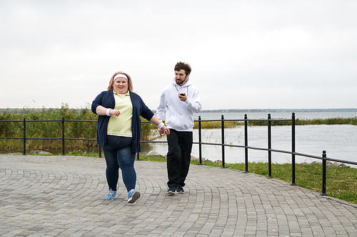 Full length portrait of overweight woman running outdoors with personal trainer, copy space