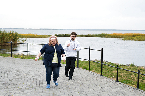 Full length portrait of obese woman running outdoors with personal trainer motivating her, copy space