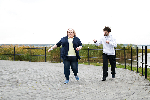 Full length portrait of obese young woman running outdoors with personal trainer motivating her, copy space