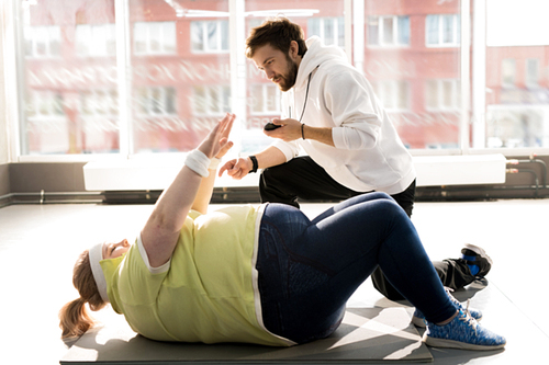 Side view portrait of obese young woman doing crunches during workout with fitness instructor in sunlight, copy space