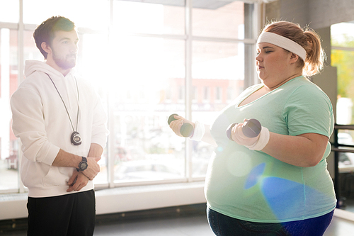 Waist up portrait of obese young woman working out with dumbbells during weight loss training with fitness instructor in sunlight