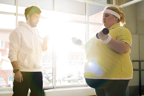 Waist up portrait of overweight young woman working out with dumbbells during weight loss training with fitness instructor in sunlight