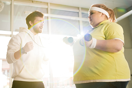 Waist up portrait of fat young woman working out with dumbbells during weight loss training with fitness instructor in sunlight