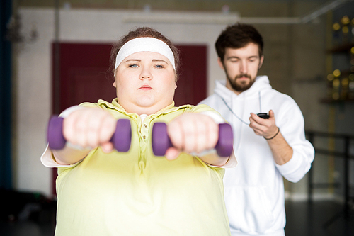 Front view portrait of motivated overweight woman working out with dumbbells during weight loss training with fitness instructor