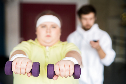 Front view portrait of motivated overweight woman working out with dumbbells during weight loss training with fitness instructor, focus on foreground