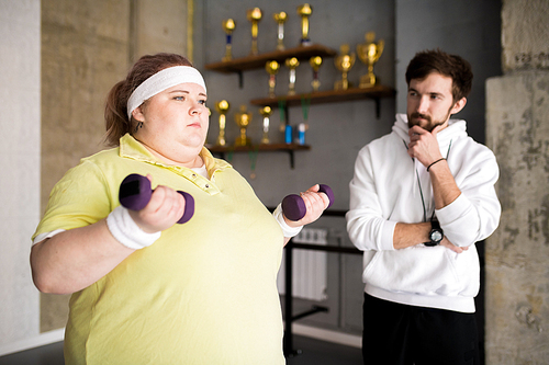 Portrait of motivated overweight woman working out with dumbbells during weight loss training with fitness instructor, copy space