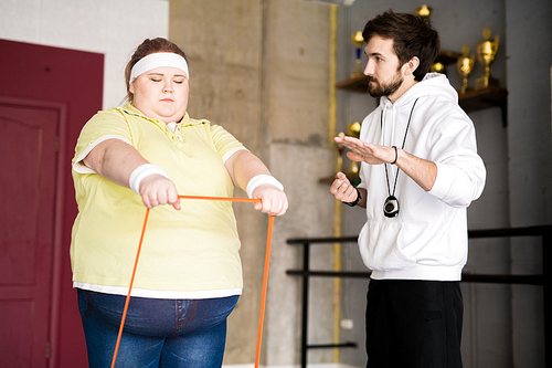 Waist up portrait of obese young woman working out using elastic band with personal fitness instructor in health club, copy space