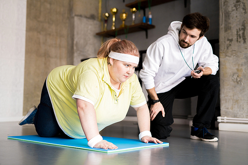 Portrait of obese young woman working out on floor with personal fitness instructor in health club, copy space