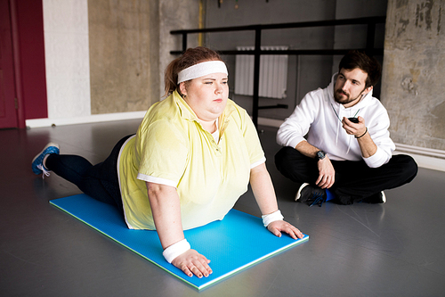 Full length portrait of  overweight young woman working out on floor with personal fitness instructor in health club, copy space