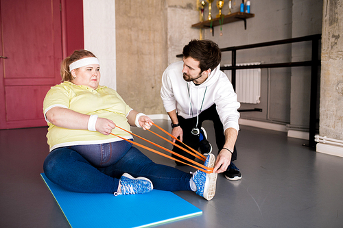 Full length portrait of  overweight woman working out on floor with personal fitness instructor in health club, copy space