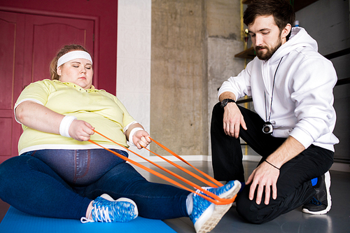 Full length portrait of  overweight woman stretching on floor with personal fitness instructor in health club, copy space