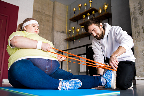 Low angle  portrait of  overweight woman stretching on floor with personal fitness instructor in health club, copy space