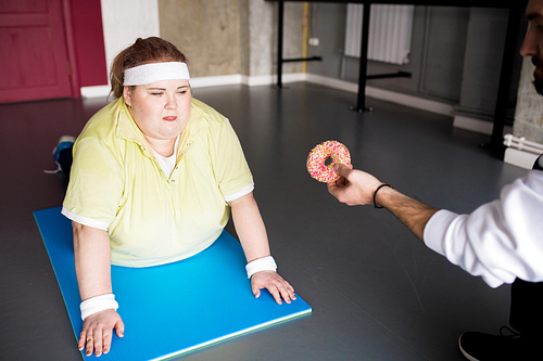 Portrait of obese young woman working out lying on mat and looking at donut, copy space