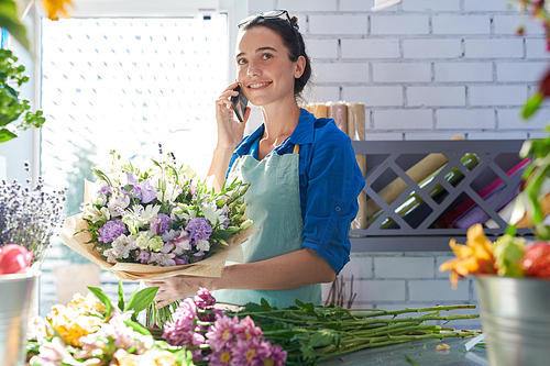 Young smiling florist holding flowers and connecting on mobile phone near workplace