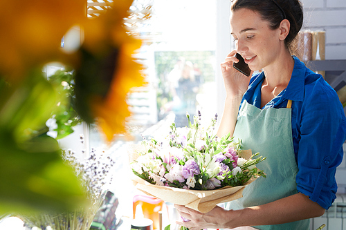 Side view portrait of smiling female florist speaking by phone while arranging flowers in sunlit shop, copy space