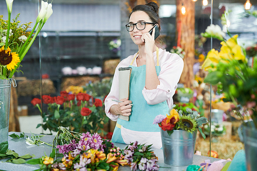 Waist up portrait of smiling female shopkeeper speaking by phone while arranging flowers in small flower shop, copy space