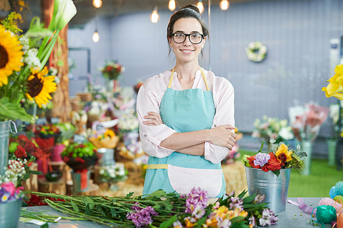 Waist up portrait of smiling female shopkeeper posing with arms crossed and   in small flower shop, copy space