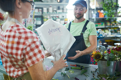Portrait of smiling delivery guy taking wrapped bouquet from shopkeeper in flower shop, copy space