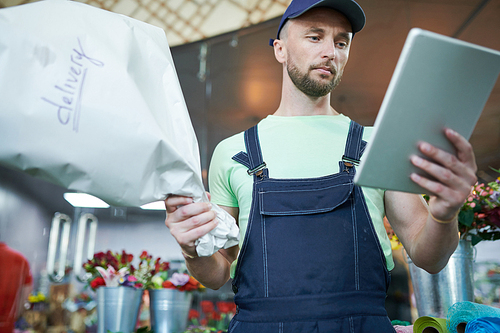 Low angle portrait of handsome delivery guy holding digital tablet and bouquet checking order info in flower shop