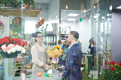Wide angle portrait of smiling young woman giving bouquet of beautiful flowers to customer in small flower shop, copy space