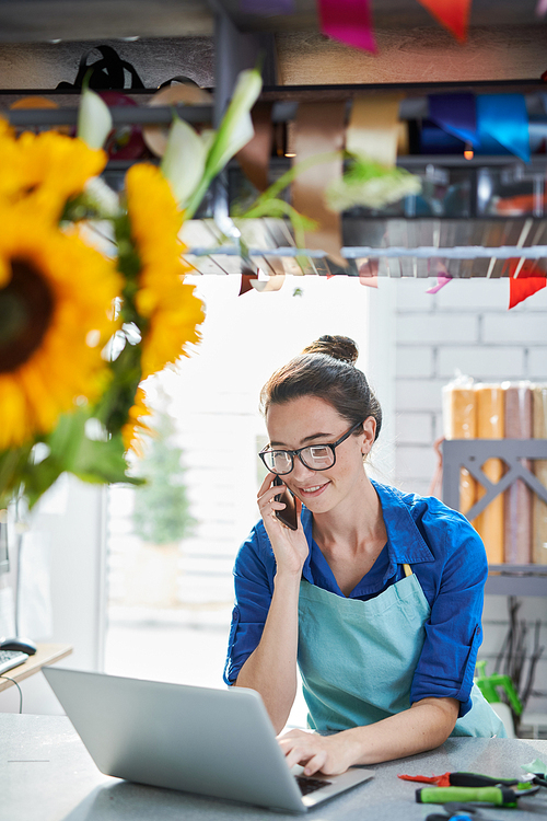 Portrait of modern young businesswoman speaking by phone and using laptop at counter in flower shop, copy space