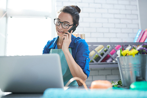 Waist up portrait of modern young businesswoman speaking by phone and using laptop at counter in flower shop, copy space