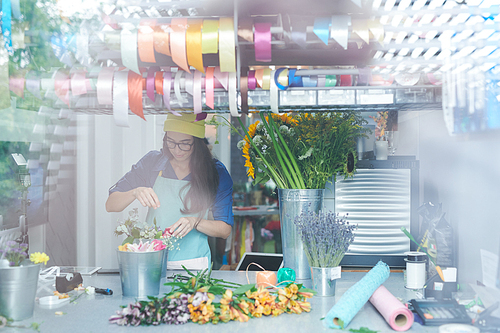 Portrait of contemporary young woman arranging flower compositions while working in shop, shot from behind glass wall