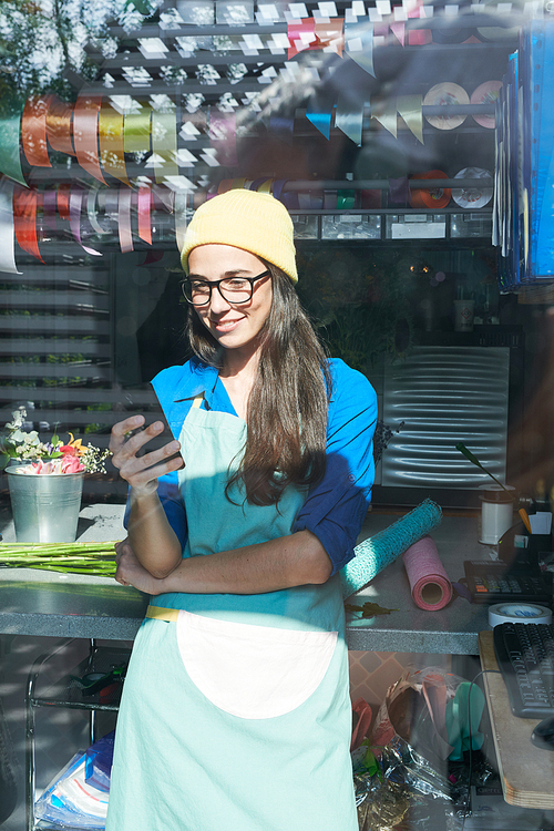 Portrait of creative young woman wearing apron using phone and smiling while standing in sunlight in flower shop