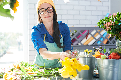 Portrait of contemporary young woman wearing glasses arranging flower compositions and smiling happily while enjoying work in sunlit shop, copy space