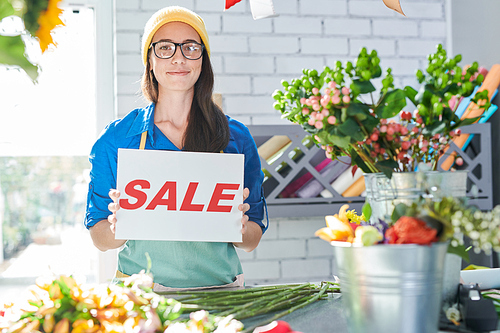 Portrait of happy contemporary woman posing in flower shop holding red SALE sign, copy space